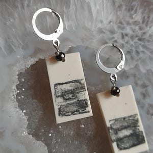 May include: A pair of rectangular earrings with a cream-colored base and a black, textured design. The earrings have silver-colored lever-back closures and a small black bead. The earrings are displayed on a light gray surface.