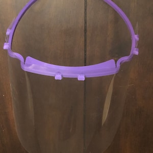 May include: A clear plastic face shield with a purple headband. The headband has two loops on the top and is adjustable.