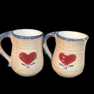 May include: Two ceramic mugs with a beige speckled finish. Each mug features a red heart design with blue accents and a blue rim and handle. The mugs are set against a black background.