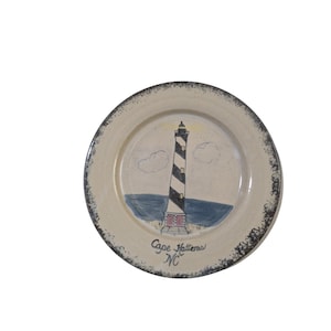 Peut inclure: Une assiette ronde en céramique avec une finition crème mouchetée et un bord texturé foncé. Le centre présente un phare en spirale noir et blanc peint à la main, un océan bleu et deux nuages. Le texte Cape Hatteras NC est écrit en noir.