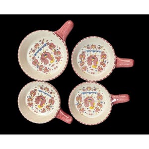 以下が含まれることがあります： 取っ手付きのセラミックボウル4つ。それぞれ手描きのデザインが施されています。ボウルはクリーム色で、赤と黄色の雄鶏のモチーフが描かれています。取っ手と縁は赤一色です。ボウルは黒い背景に配置されています。