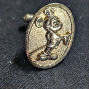 May include: Gold-toned cufflink featuring a raised image of Mickey Mouse in a classic pose. The oval-shaped cufflink has a textured background and a post for attachment. A vintage-style accessory.
