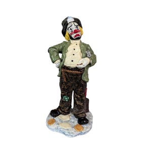May include: A hand-painted ceramic figurine of a clown with a green jacket, brown pants, and a white shirt. The clown is holding a few coins in his hand and has a sad expression on his face. The figurine is standing on a base with a stone-like texture.