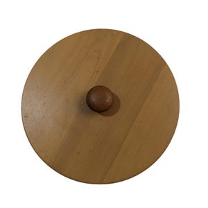 May include: A round, light brown wooden lid with a small, dark brown knob in the center. The lid has a smooth surface and a natural wood grain pattern. The knob is a rounded sphere.