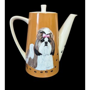 May include: A ceramic teapot with a tan body and white handle and spout. The teapot features a hand-painted Shih Tzu dog with a pink bow. Black paw prints decorate the bottom of the teapot.