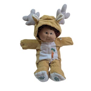 May include: A Cabbage Patch Kids doll wearing a tan and white deer costume with a hood and antlers. The doll has brown hair and a neutral expression. The costume has a carrot and tree design on the front.