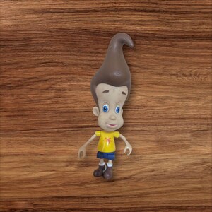 May include: A Jimmy Neutron action figure. The figure has a large, brown, cone-shaped hairstyle, blue eyes, and a yellow shirt with a flower design. He is wearing blue shorts, brown shoes, and white socks. The figure is standing on a wooden surface.