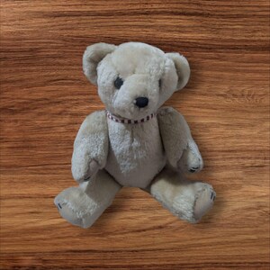 May include: A beige teddy bear with a red and white striped collar. The bear has black eyes and a black nose. The bear is sitting with its legs spread apart. The bear is made of a soft, plush material.