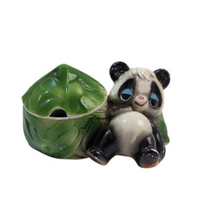 May include: A ceramic panda figurine attached to a green, leaf-shaped container. The panda has black and white fur, blue eyelids, and a small pink nose. The container features a textured, glossy green surface with a lid that resembles a large leaf.