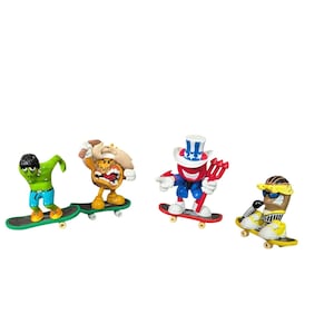 May include: Four novelty figurines on skateboards. The figures include a green zombie, a burger, Uncle Sam, and a mummy. Each figure is on a skateboard with wheels. The figures are brightly colored and detailed.