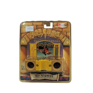 May include: A vintage Harry Potter and the Sorcerer's Stone 3D Viewer toy, packaged in a blister pack. The viewer is yellow and black, with the Harry Potter logo and 3D Viewer text visible. The packaging features a brick design.