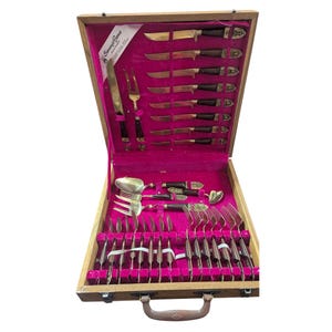 May include: A vintage silverware set in a wooden case with a pink velvet interior. The set includes knives, forks, spoons, and serving utensils. The knives have brass blades and dark brown handles. The case is open, revealing the neatly arranged pieces.