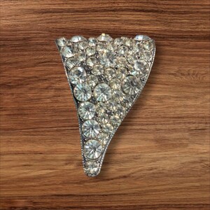 May include: A silver-toned, triangular brooch encrusted with numerous clear, sparkling rhinestones. The brooch has a curved bottom edge and a pointed top, set against a warm, brown wooden surface.