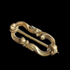 May include: A gold-colored brooch with an ornate, openwork design. The brooch features a central bar and decorative, swirling elements. The pin is set against a black background, highlighting the intricate details of the jewelry.