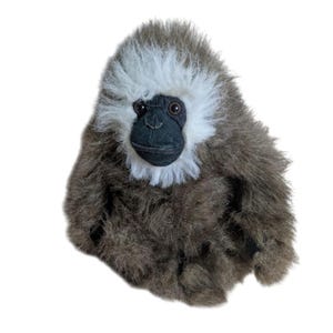 May include: A plush toy of a brown and white gibbon monkey with black eyes and a black nose. The monkey is sitting with its legs crossed and its arms folded.