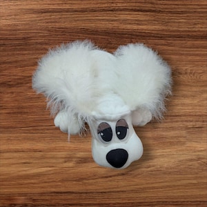May include: A white plush dog toy with large, fluffy ears and a black nose. The dog has large, expressive eyes with long eyelashes. The toy is lying on a wooden surface.