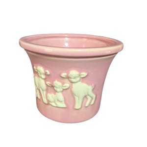 May include: A pink ceramic planter with a flared shape. The planter features a raised relief design of three white lambs. The rim of the planter is slightly lighter pink than the body.