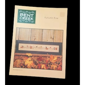 May include: A craft pattern by Bent Creek, titled "Autumn Row". The cover displays a cross-stitch design with pumpkins and autumn leaves. The design includes the alphabet and is set against a wooden backdrop.