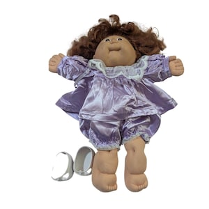 May include: A vintage doll with brown curly hair, wearing a lavender satin outfit with lace trim. The doll has a round face and chubby limbs. White shoes are placed near the doll.
