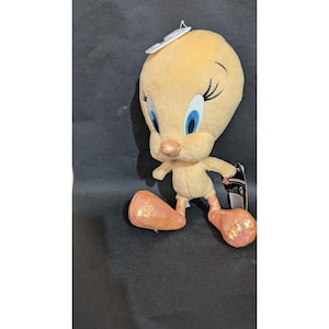 May include: A plush Tweety Bird toy in yellow with blue eyes, a pink nose, and orange feet. The toy has a white tag attached to its head. The plush toy is a character from the Looney Tunes cartoon series.