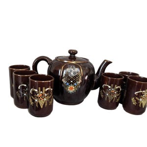 May include: A brown ceramic tea set with a teapot and six matching cups. The teapot and cups feature a decorative floral and geometric design with gold accents. The set is designed for serving tea.