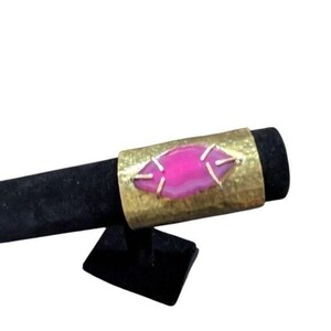 May include: A gold-toned metal ring with a hammered texture. The ring features a vibrant pink, oval-shaped stone at the center, secured with gold-colored accents. The ring is displayed on a black stand, showcasing its design and color.