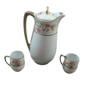 May include: A white porcelain tea set with a floral design. The set includes a tall pitcher with a lid and two small cups. The pitcher and cups have gold trim and handles. The floral design features pink roses.