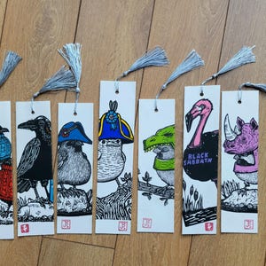 Può includere: Sette segnalibri rettangolari in carta con nappe argentate, ognuno caratterizzato da illustrazioni a inchiostro bianco e nero di uccelli che indossano accessori colorati, tra cui maglioni, cappelli e maschere di animali, con piccoli timbri rossi.