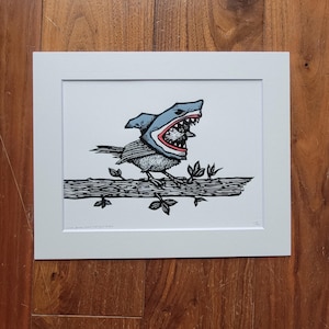 Puede incluir: Una impresión en blanco y negro enmarcada de un pájaro con un sombrero de tiburón, posado en una rama con hojas. El pájaro está detallado con sombreado texturizado. El sombrero de tiburón es azul y blanco con un borde rojo.