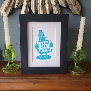 May include: A framed print featuring a blue and white illustration of a bird wearing a hat and holding a sign with the text "It's a bit of a kerfuffle." The print is in a black frame, with green glass candle holders and white candles.