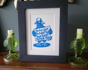 Op de afbeelding: Een ingelijste print met een blauwe illustratie van een vogel met een hoed op, die een bord vasthoudt met de tekst "You've got to fight for your right to pour tea". Het kunstwerk zit in een donkerhouten lijst met een witte passe-partout, geflankeerd door twee groene, visvormige glazen kandelaars.