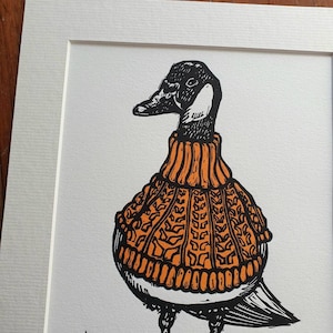 K&ouml;nnte beinhalten: Eine Schwarzwei&szlig;illustration einer Gans, die einen orangefarbenen Strickpullover tr&auml;gt. Die Gans steht auf einer braunen Oberfl&auml;che mit gr&uuml;nem Gras.