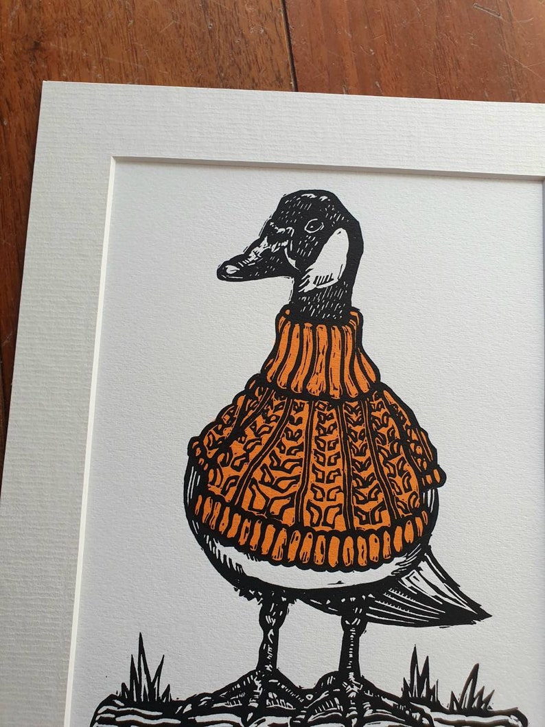 K&ouml;nnte beinhalten: Eine Schwarzwei&szlig;illustration einer Gans, die einen orangefarbenen Strickpullover tr&auml;gt. Die Gans steht auf einer braunen Oberfl&auml;che mit gr&uuml;nem Gras.