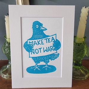 Pode incluir: Uma impressão de arte emoldurada com um pombo azul segurando uma placa que diz "MAKE TEA NOT WAR". A obra de arte é definida contra um fundo branco e emoldurada em um passe-partout branco. A impressão é exibida em uma superfície de madeira.
