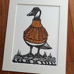 K&ouml;nnte beinhalten: Eine Schwarzwei&szlig;illustration einer Gans, die einen orangefarbenen Strickpullover tr&auml;gt. Die Gans steht auf einem Baumstamm mit Steinen und Gras.