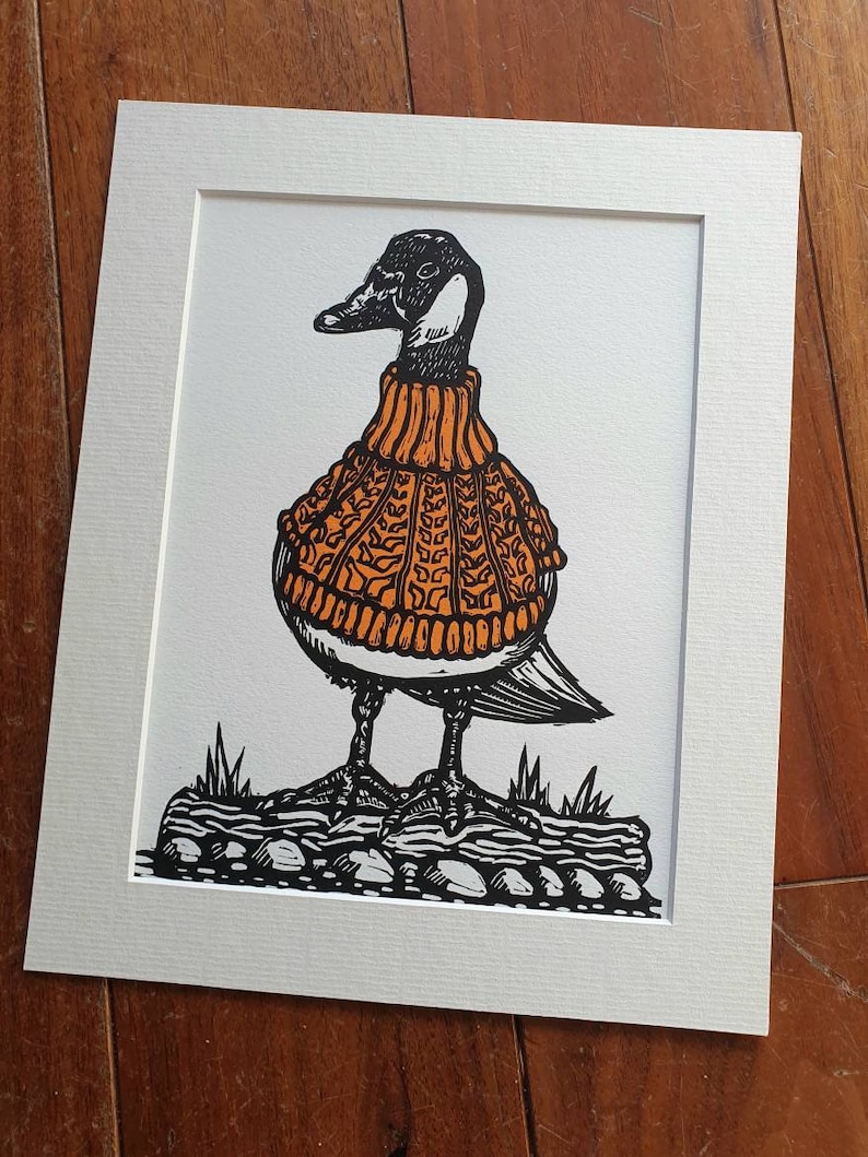 K&ouml;nnte beinhalten: Eine Schwarzwei&szlig;illustration einer Gans, die einen orangefarbenen Strickpullover tr&auml;gt. Die Gans steht auf einem Baumstamm mit Steinen und Gras.