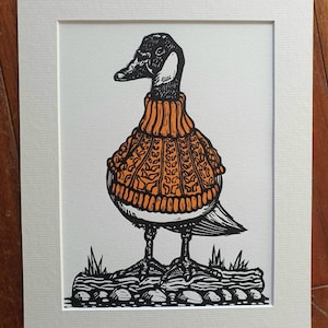 K&ouml;nnte beinhalten: Eine Schwarzwei&szlig;illustration einer Gans, die einen orangefarbenen Strickpullover tr&auml;gt. Die Gans steht auf einem Stein mit Gras im Hintergrund.