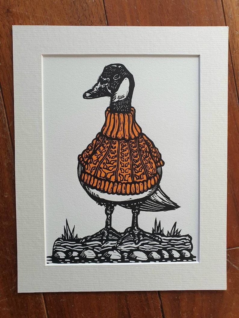 K&ouml;nnte beinhalten: Eine Schwarzwei&szlig;illustration einer Gans, die einen orangefarbenen Strickpullover tr&auml;gt. Die Gans steht auf einem Stein mit Gras im Hintergrund.