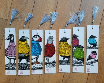 Puede incluir: Siete marcapáginas rectangulares de papel con ilustraciones de aves vistiendo jerséis de punto coloridos. Cada marcapáginas tiene una borla plateada en la parte superior. Las aves incluyen un ganso, cuervo, frailecillo, paloma y otras figuras.
