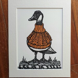 K&ouml;nnte beinhalten: Eine Schwarzwei&szlig;illustration einer Gans, die einen orangefarbenen Strickpullover tr&auml;gt. Die Gans steht auf einem felsigen Untergrund.