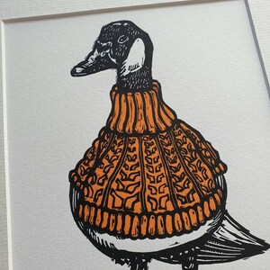 K&ouml;nnte beinhalten: Eine Schwarzwei&szlig;illustration einer Gans, die einen orangefarbenen Strickpullover tr&auml;gt. Die Gans steht auf einer gr&uuml;nen Grasfl&auml;che.