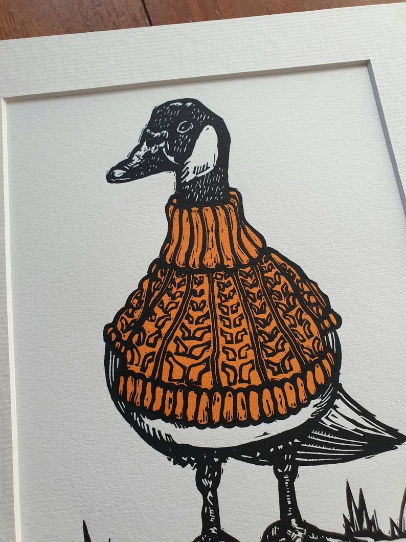 K&ouml;nnte beinhalten: Eine Schwarzwei&szlig;illustration einer Gans, die einen orangefarbenen Strickpullover tr&auml;gt. Die Gans steht auf einer gr&uuml;nen Grasfl&auml;che.