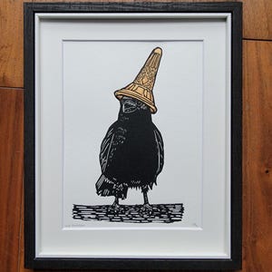 May include: Framed black and white art print featuring a crow wearing a gold ice cream cone hat. The crow stands on a wooden surface. The artwork is set against a white background.