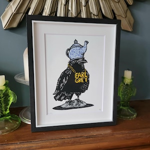 Könnte beinhalten: Ein gerahmter Druck mit der Illustration eines schwarzen Raben, der eine hellblaue, gemusterte Teekanne auf dem Kopf trägt und eine goldene Kette mit einem gelben Anhänger, auf dem in schwarzen Buchstaben EARL GREY steht. Das Bild ist in einem schwarzen Rahmen mit weißem Passepartout.