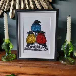 Puede incluir: Una impresión enmarcada de tres cuervos negros que llevan suéteres de punto de colores. Los cuervos están posados en una rama con un fondo blanco.