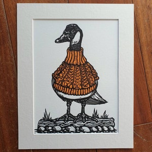 K&ouml;nnte beinhalten: Eine Schwarzwei&szlig;illustration einer Gans, die einen orangen Pullover tr&auml;gt. Die Gans steht auf einem Stein mit Gras im Hintergrund.