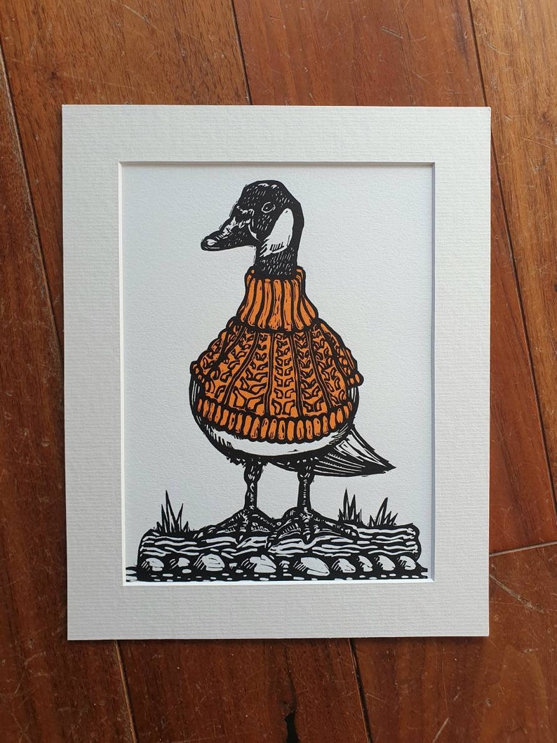 K&ouml;nnte beinhalten: Eine Schwarzwei&szlig;illustration einer Gans, die einen orangen Pullover tr&auml;gt. Die Gans steht auf einem Stein mit Gras im Hintergrund.