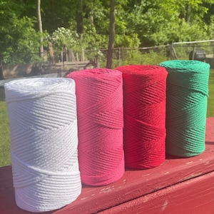 May include: Four spools of cotton rope in white, pink, red, and green. The spools are arranged in a row on a red wooden surface.