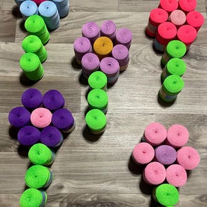 May include: Arrangement of colorful spools of thread, crafted to resemble flowers. The 'petals' are various shades of pink, purple, blue, and yellow, with green stems. The spools are arranged on a wooden surface.