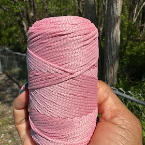 May include: A spool of pink yarn, held in a hand, with varying shades of pink. The yarn is tightly wound, showing a textured surface. The background features green trees and a chain-link fence.